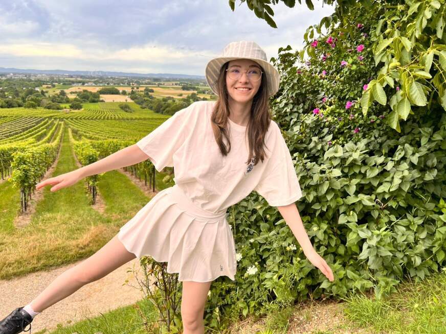 Emmi steht in der Natur, balanciert auf einem Bein und trägt einen Hut.